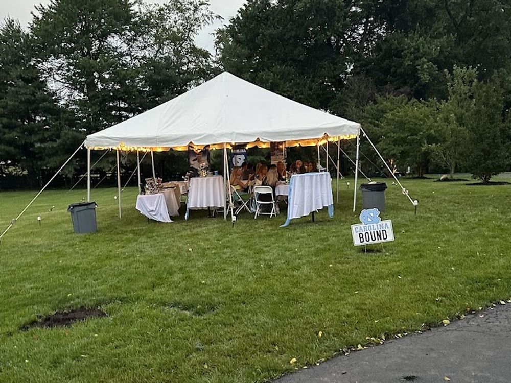 A white frame tent lit from within at dusk, set up on a back lawn for a graduation party