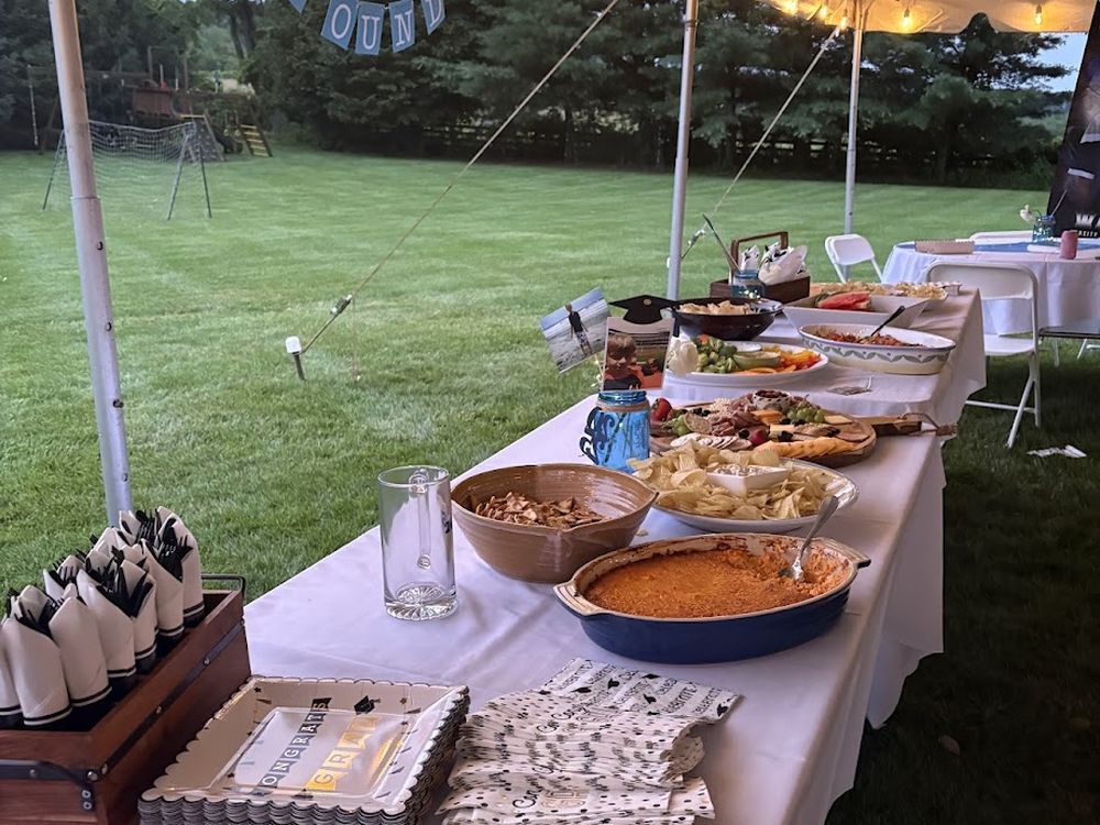 A long buffet table dressed in white linen under a tent hung with string lights at dusk